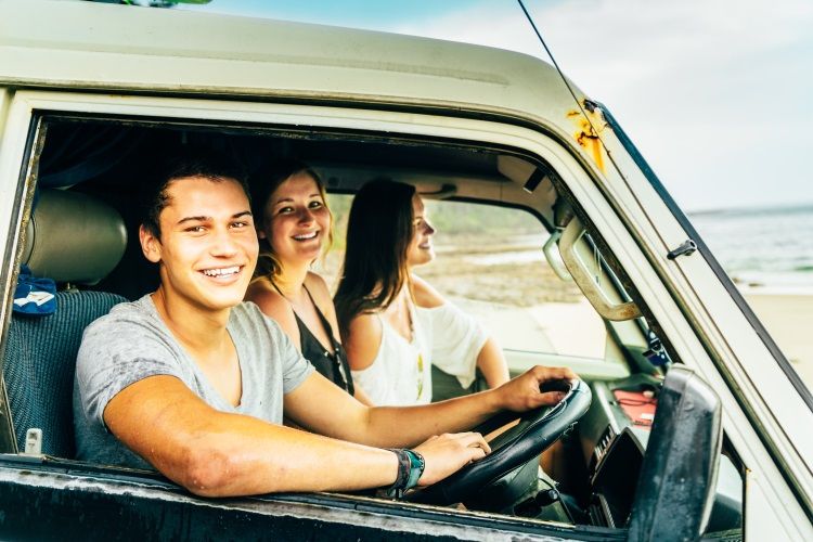 Drei Personen lachen während sie in ihrem Campervan in Australien am Strand sitzen.